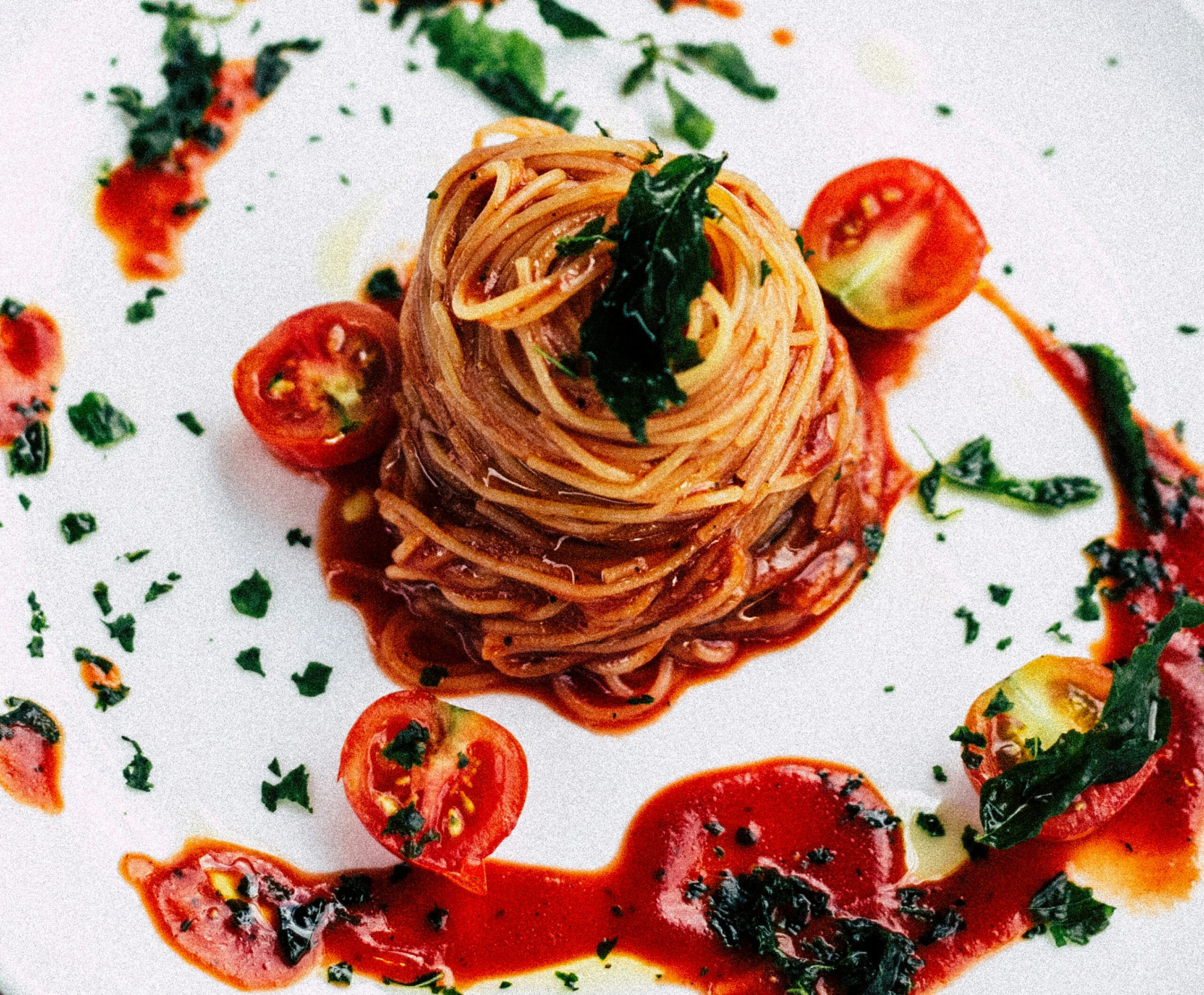 Close up of pasta arranged in a swirl on white plate with cherry tomatoes and tomato sauce with greens as garnish