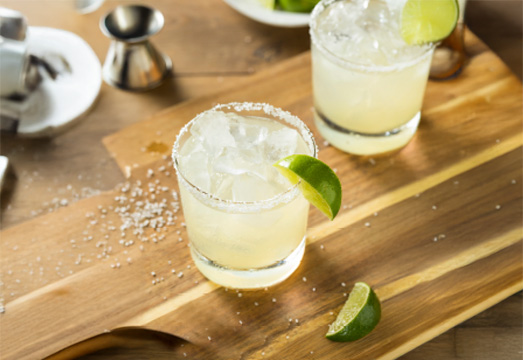 A wood table top with two rock glasses filled with prosecco margarita cocktail and lime garnish