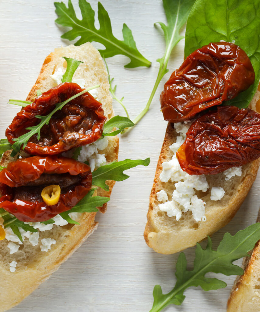 A close up of two toasted crustini with arugula, crumbled cheese, and sun dried tomatoes
