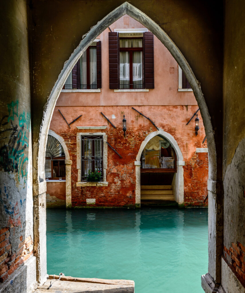 A venice scene showing a two story stucco building with arched doorways and exposed brick with a canal as seen through an arched doorway with graffiti on its walls