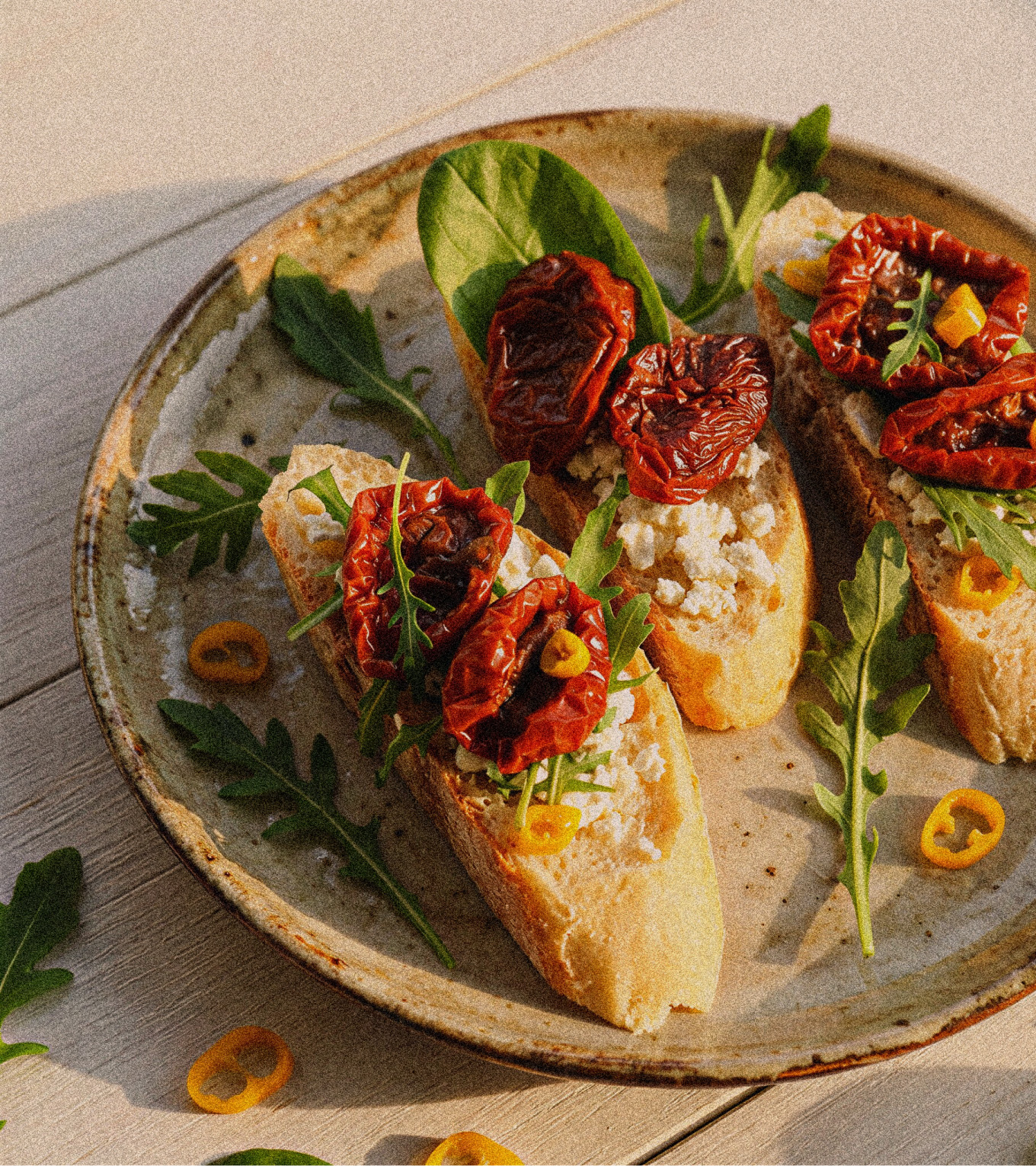 A plate of three toasted crustini with arugula, crumbled cheese, and sun dried tomatoes