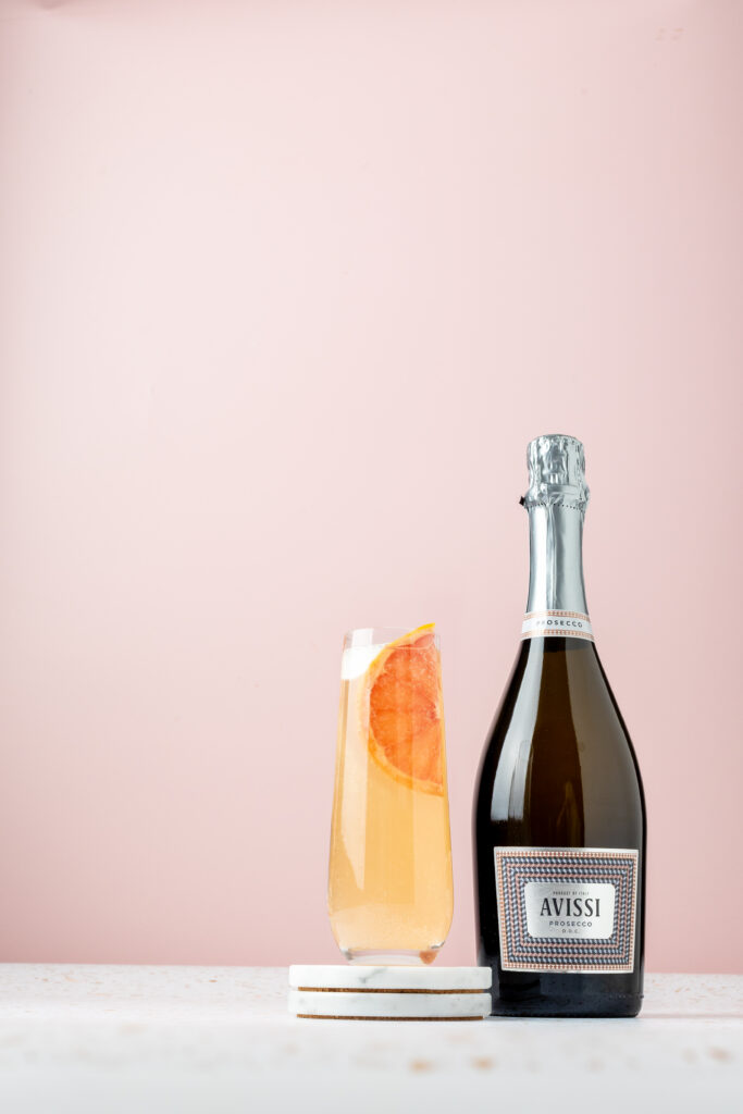 Still life of a bottle of a orange colored cocktail in a tall tapered glass on a marble coaster with a bottle of Avissi Prosecco to the right of it, on a marble surface, with a pink background
