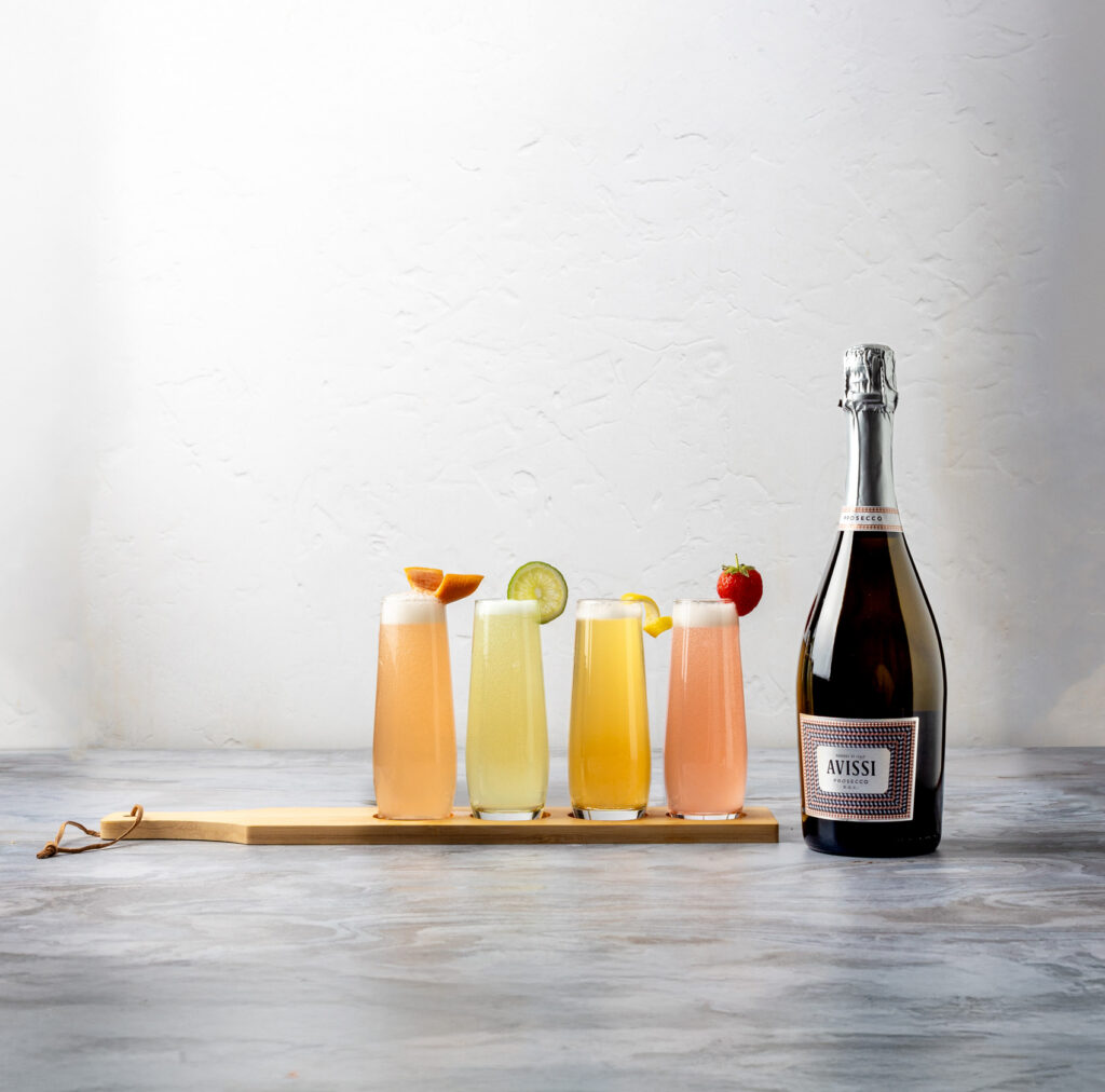 A tabletop arrangement of four stemless flutes with differently colored prosecco cocktails that each have a different kind of fruit garnish, all sitting on a wood cutting board, with a bottle of Avissi Prosecco