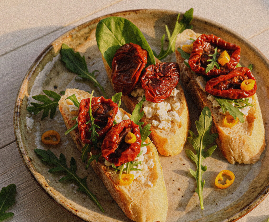 Close up of crostini with cheese, dried tomatoes, arugula, and basil, on an earthy ceramic plate