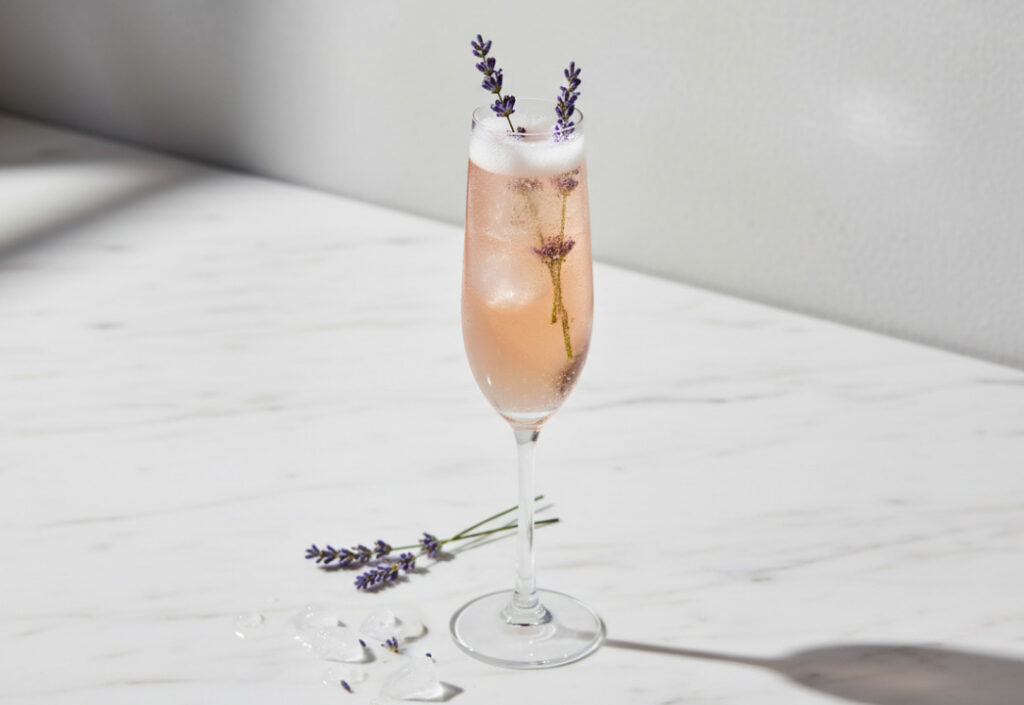 Close up of a pale pink cocktail in a champagne flute glass with purple lavender sprigs garnish, on a marble surface