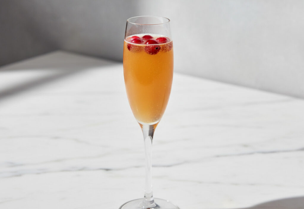 Close up of an orange colored cocktail in a champagne flute glass with red berries floating on top, on a marble surface
